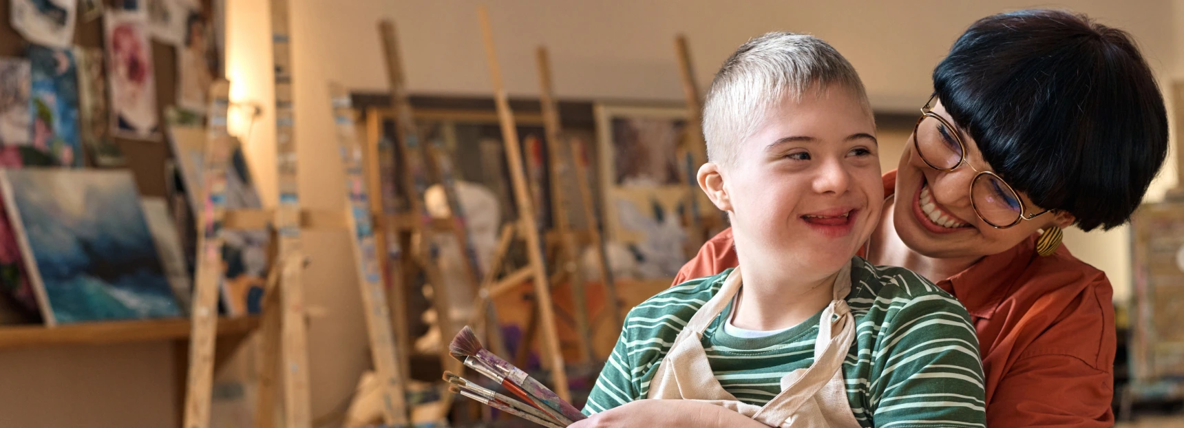 Portrait of smiling young woman embracing boy with disability while enjoying art class in studio copy space