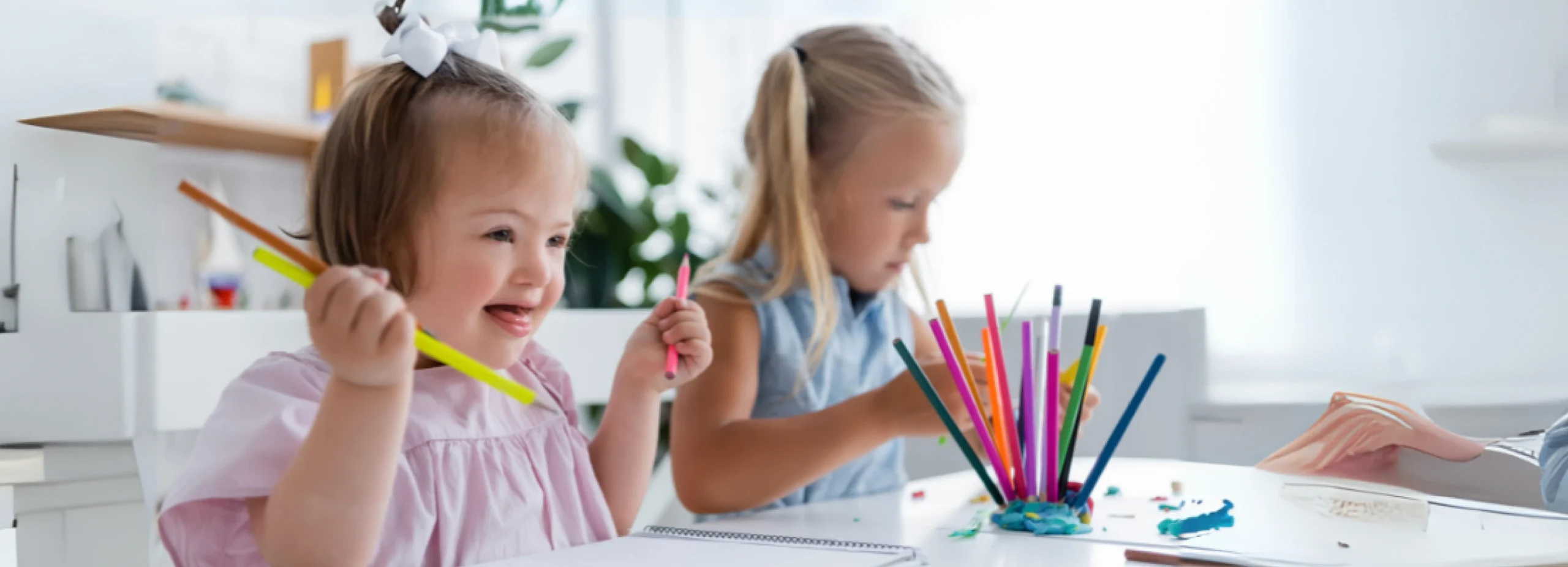 happy toddler kid with down syndrome holding pencils near blurred blonde girl